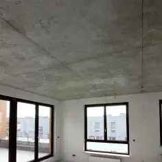 Surowy betonowy sufit z widocznymi liniami podziału i oknami wychodzącymi na budynki.