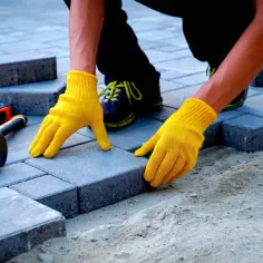 Ręce w żółtych rękawiczkach układają kostkę brukową. Zastanawiasz się, ile kosztuje układanie kostki brukowej? Narzędzia leżą obok.