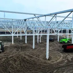 Budowa hali produkcyjnej: szkielet konstrukcji stalowej, ciągniki, koparka i wywrotka na placu budowy.