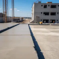 Świeżo wylany beton na placu budowy, gdzie pracownicy obawiają się przymrozku. W tle budynek w stanie surowym i dźwigi.