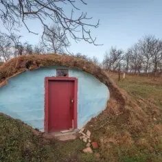 Gotowa piwniczka betonowa, częściowo zasypana ziemią i trawą, z niebieską ścianą i czerwonymi drzwiami.