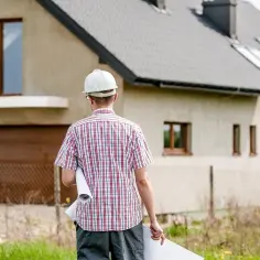Inżynier z planami w ręku przed domem jednorodzinnym. Zbliża się zawiadomienie o zakończeniu budowy.