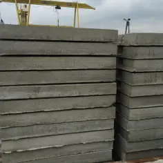Stos grubych płyt drogowych betonowych czeka na ułożenie. W tle żółty dźwig budowlany.