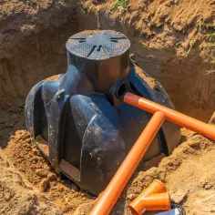 Instalacja szamba betonowego w wykopie. Pomarańczowe rury kanalizacyjne i poziomica wskazują na trwające prace.