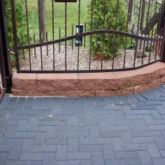 Nowoczesna ścieżka z ciemnej kostki brukowej, ozdobny murek i kuta brama. Zastanawiasz się, ile za metr kostki brukowej?