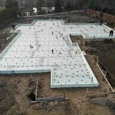 Budowa fundamentów: biała folia pod płytę fundamentową, zbrojenie i rury. Dwa pracownicy przygotowują teren pod wylanie betonu.