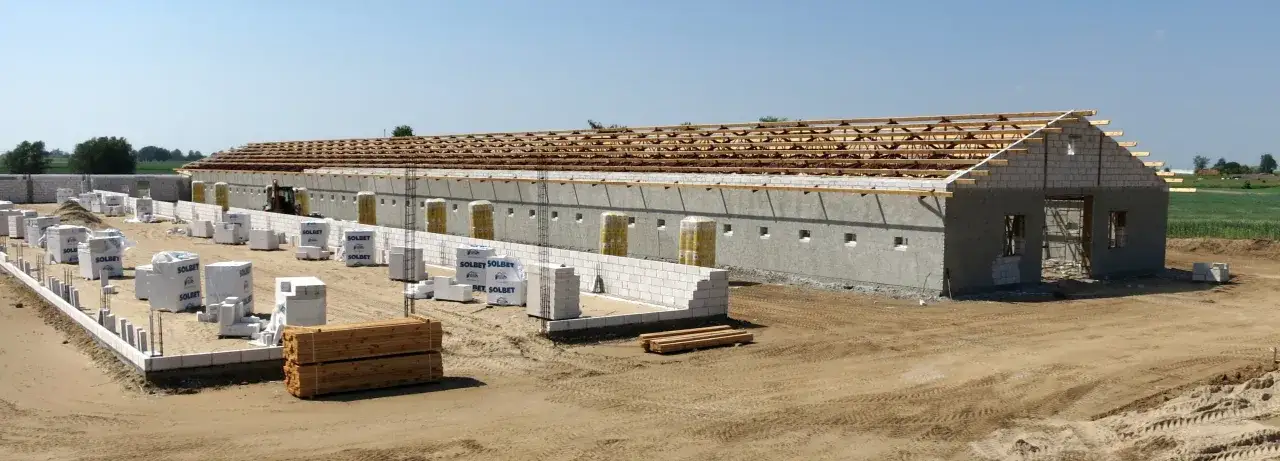 Budowa kurnika z płyt betonowych w toku. Widać materiały budowlane, drewniane elementy konstrukcji dachu i gołe ściany.