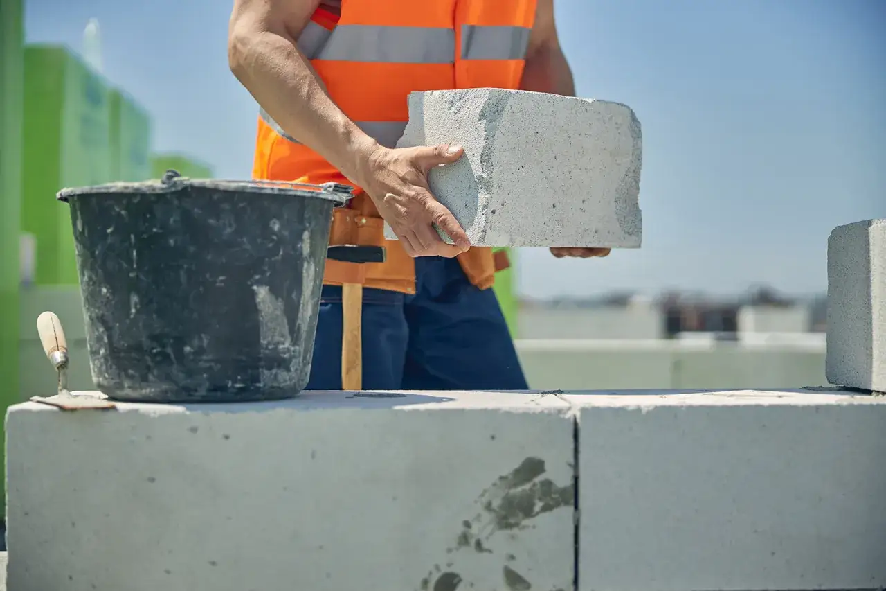 Budowlaniec w kamizelce odblaskowej układa bloczek betonowy. Dowiadujesz się, ile za murowanie z bloczka betonowego.