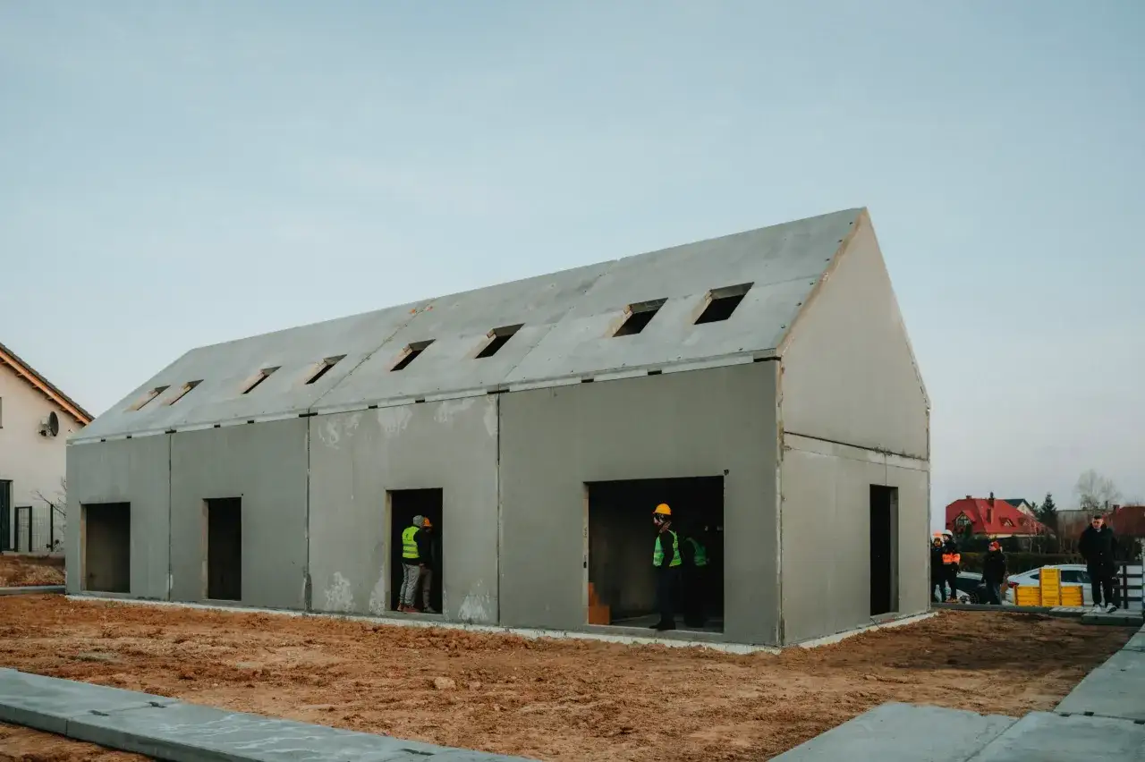 Budowa domu prefabrykowanego betonowego. Widać pracowników w kamizelkach odblaskowych i hełmach.