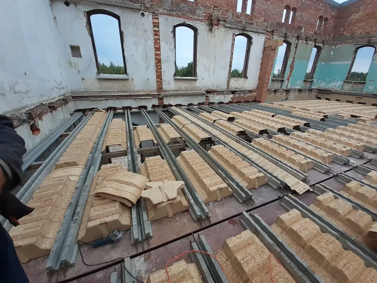 Budowa stropu na belkach stalowych w zrujnowanym budynku. Widać betonowe elementy i materiały izolacyjne.