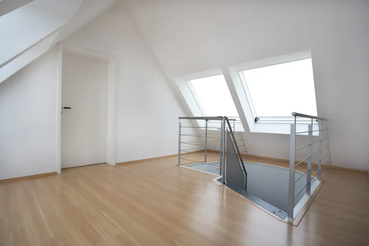 Nowoczesne schody na poddasze z metalową balustradą prowadzą w dół. Jasne okna dachowe doświetlają przestrzeń.