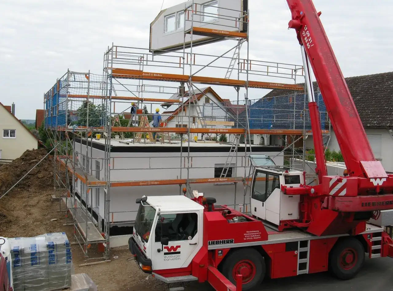 Montaż domu prefabrykowanego betonowego. Dźwig podnosi element dachu, pracownicy na rusztowaniach.