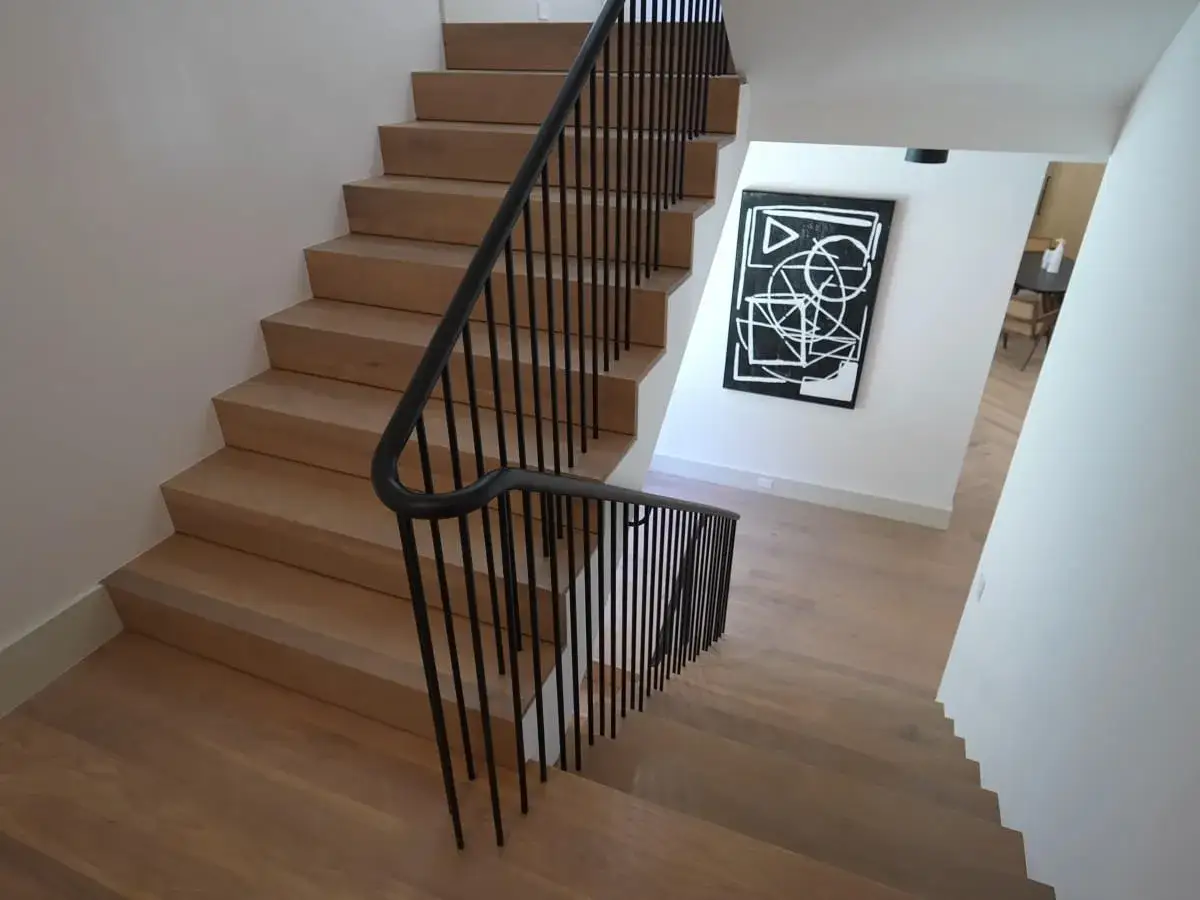Nowoczesna klatka schodowa w domu z drewnianymi stopniami i czarną, minimalistyczną balustradą. Obok wisi abstrakcyjny obraz.