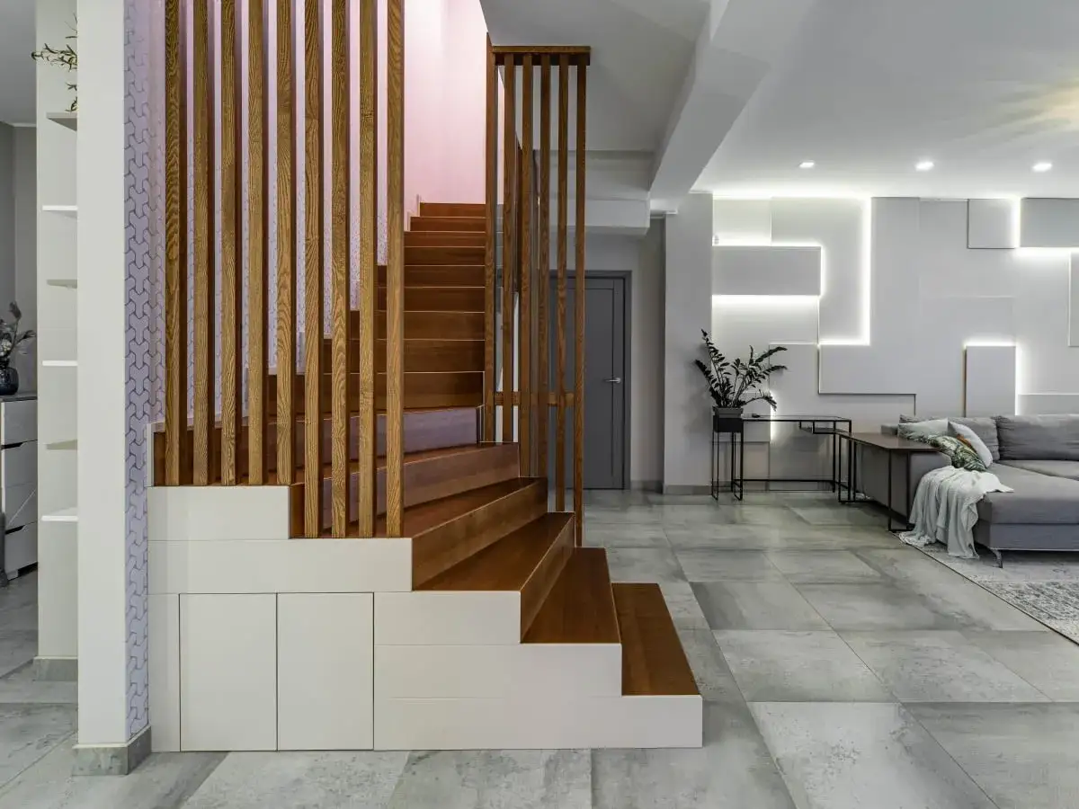 Nowoczesna klatka schodowa w domu piętrowym z drewnianymi stopniami i ażurową balustradą. Obok salon z szarą kanapą i designerską ścianą.